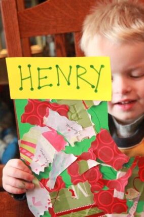 Toddler holding a paper stocking decorated with wrapping paper scraps and a name tag