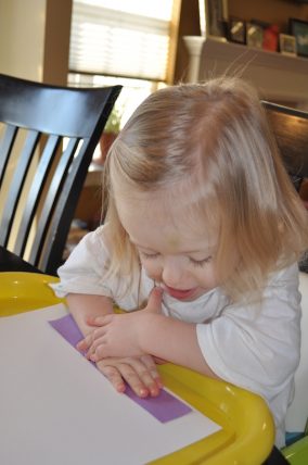 Toddler pressing a purple paper strip onto white paper to start a shape menorah craft