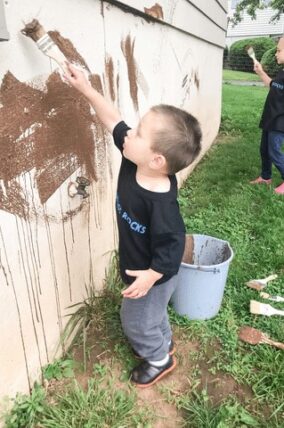 Boy painting a wall with mud using a wide brush, bucket beside him