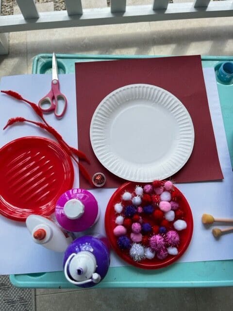 Supplies needed to make a heart snail craft with paper plates and pom poms at home.