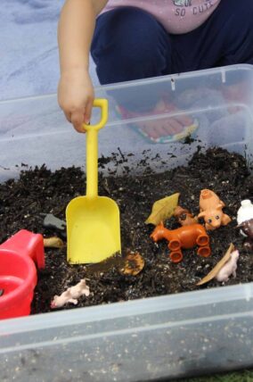 Child's hand digging with a yellow shovel in a bin of mud with toy animals