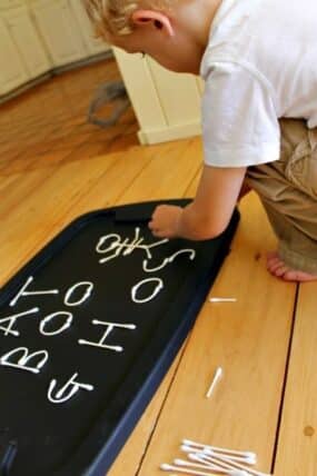 Child kneeling at a black tray writing letters with cotton swabs, a pile of swabs on the floor