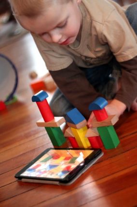 Child building a tower of wooden blocks on the floor beside a tablet