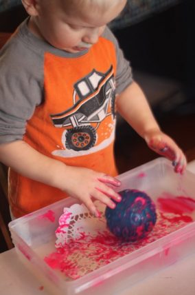 Toddler rolling a textured ball through pink paint in a plastic bin