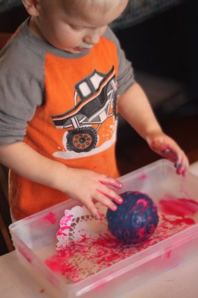 Toddler rolling a textured ball through pink paint in a plastic bin