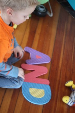 Child kneeling beside large foam letters spread across a wood floor
